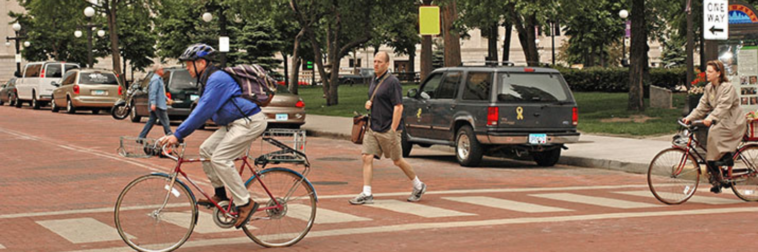 Two bicyclists and two walkers cross a brick road lined with cars.