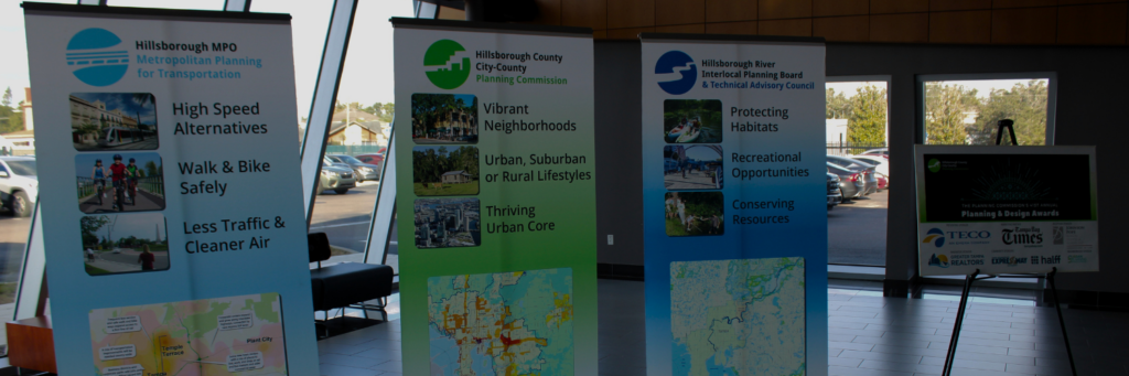 Three banners detailing the Hillsborough MPO (Metropolitan Planning for Transportation, High Speed Alternatives, walk and bike safely, less traffic and cleaner air), Hillsborough County City-County Planning Commission (vibrant neighborhoods, urban, suburban or rural lifestyles, thriving urban core), and the Hillsborough River Interlocal Planning Board and Technical Advisory Council (protecting habitats, recreational opportunities, conserving resources) stand side by side in an open, windowed lobby